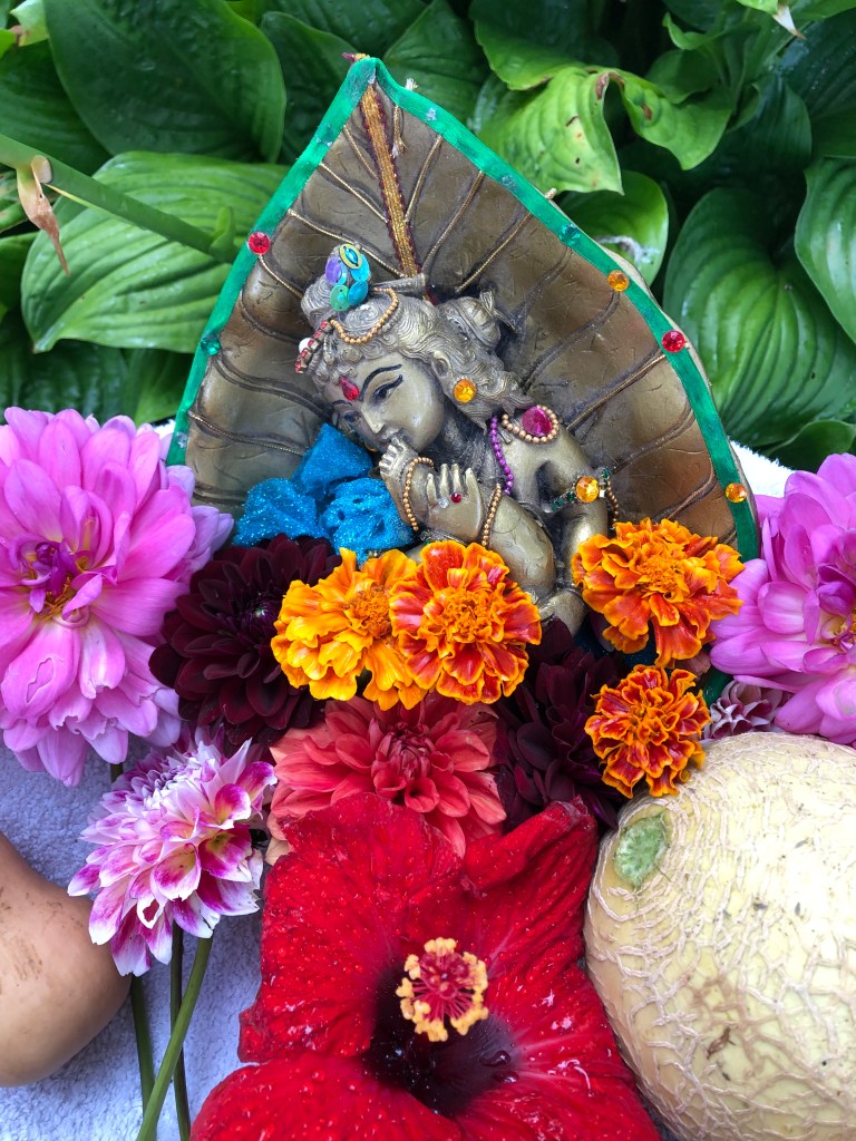 A decorative idol of a Hindu deity surrounded by colorful flowers including dahlias, marigolds, and hibiscus, set against a backdrop of lush green leaves.